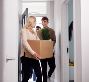 alt= "Eine Frau und zwei Männer betreten eine Wohnung. Die Frau trägt einen Pappkarton und einer der Männer hält eine Pflanze in seiner Hand."